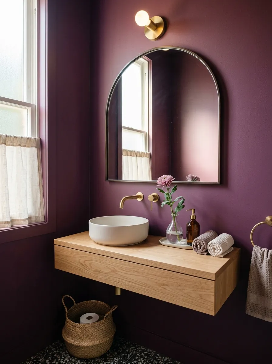 Real-life photograph of a small powder room with deep plum painted walls, oversized arched mirror, small bud vase with a single flower stem neatly placed on the counter, beautifully styled and organized, clean composition, bright natural light, vibrant plum tones, real home moody bathroom, photorealistic stock photo style.
